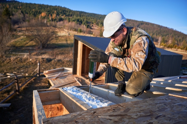 Image of a male builder applying thermal insulation on roof of wooden frame of a house